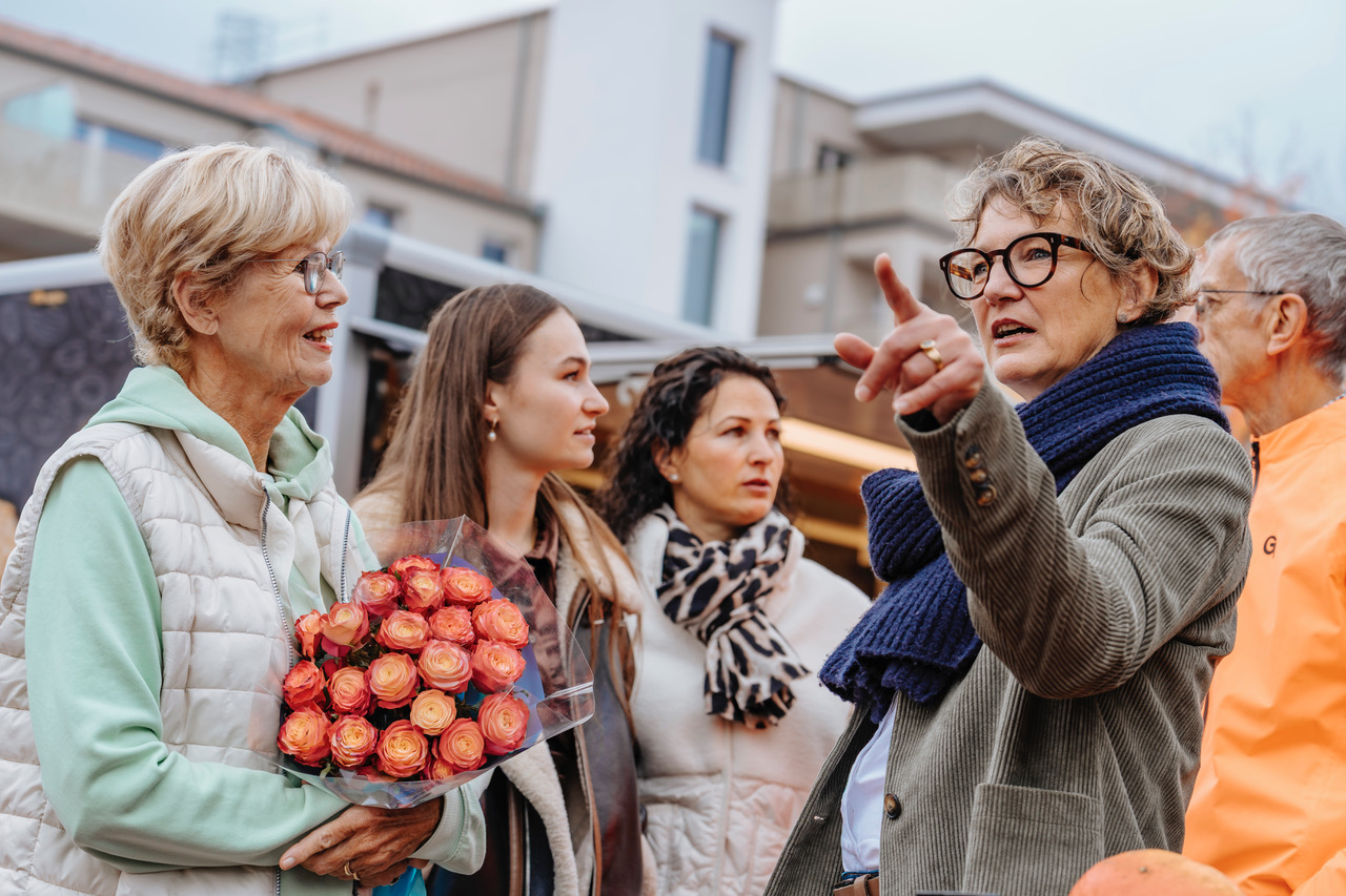 Eine ältere Frau hält bunte Blumen, während eine andere Frau auf einem belebten Markt etwas erklärt.